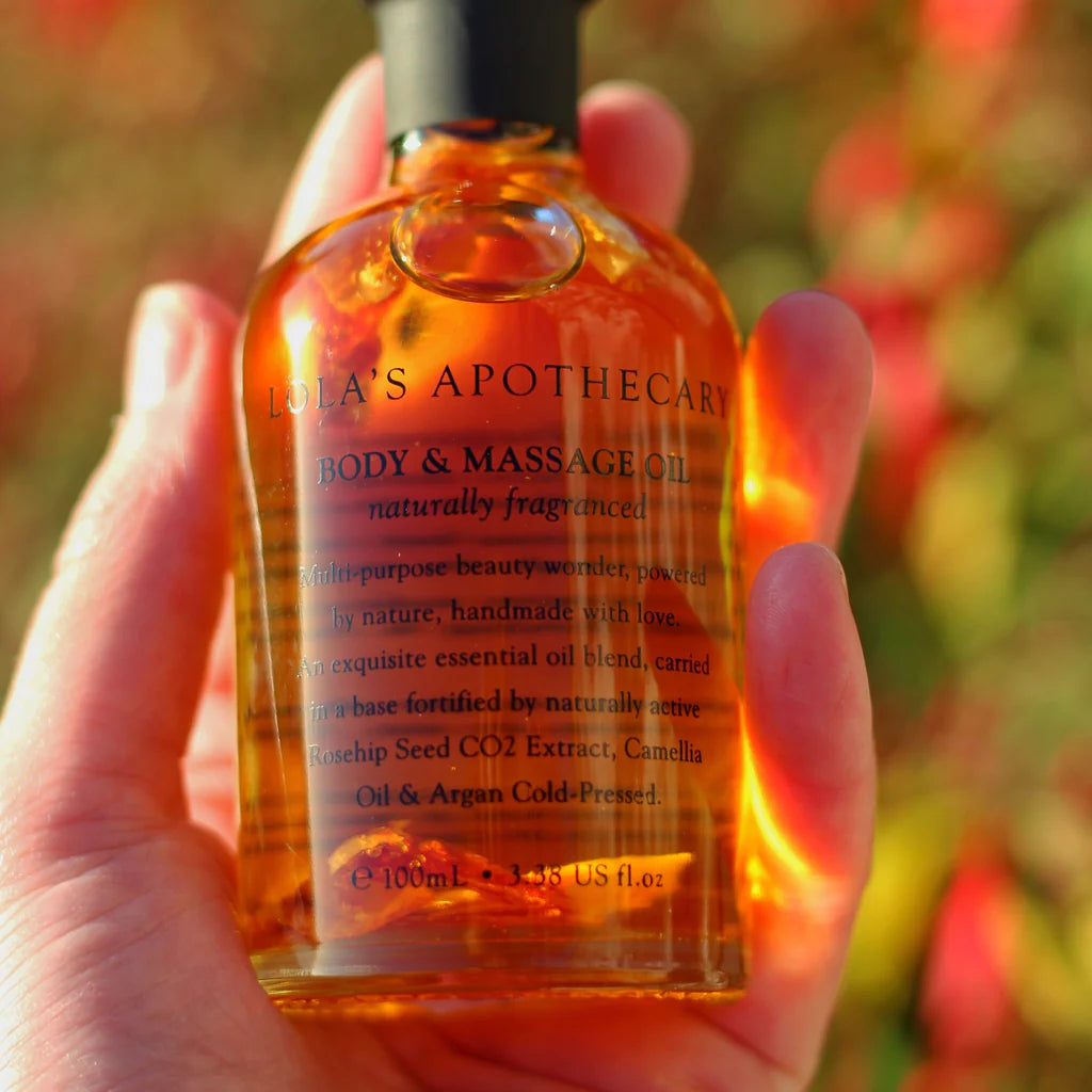 A hand holding a bottle of "euphoric indulgence" body massage oil with blurred outdoor background