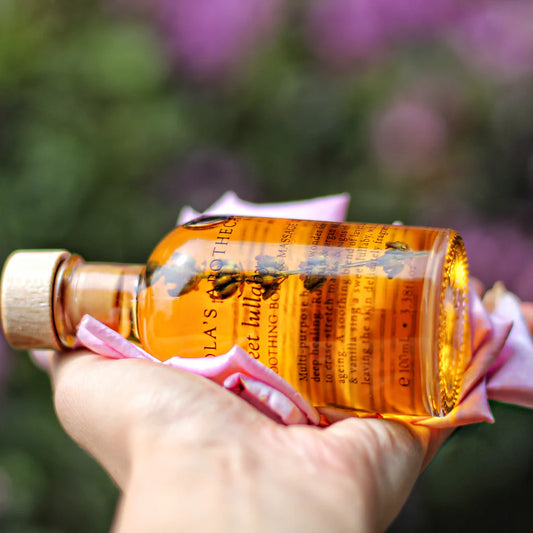 Hand holding a bottle of "Sweet Lullaby" body massage oil with blurred outdoor floral background